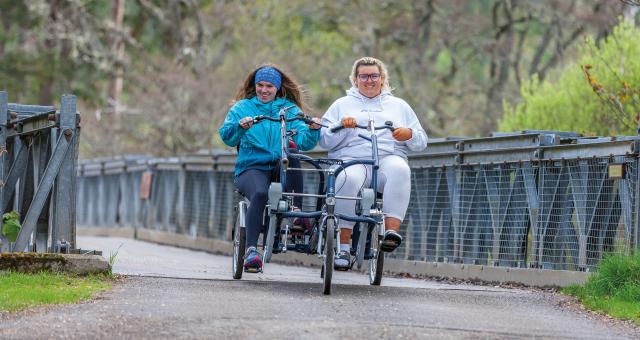 Two women are cycling a side-by-side tandem trike across a bridge. They are wearing tracksuits and are smiling