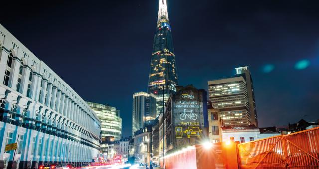 A City of London cityscape at night showing the Shard in the background. A projection is showing on a building with a cycle and a car and the words Rishi, fight climate change, don' fuel it