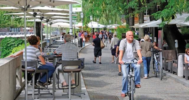 A man is cycling along a shared cycle and walking path next with restaurants on one side and people sitting at tables eating and drinking on the other