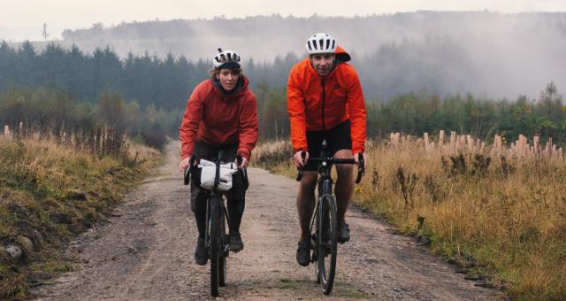 Two people are cycling along an off-road track through mountainous countryside. They are on loaded gravel bikes and wearing waterproof cycling kit