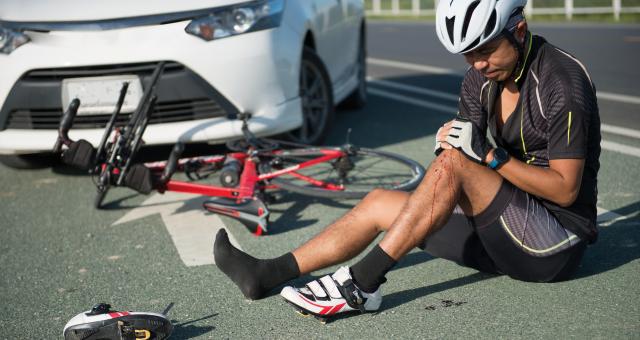 A man is sitting on the road with his legs out in front of him. One leg is bent and he's holding the knee, there is blood on his calf. He is wearing cycling kit. His bike is on the road behind him and behind that is a white car