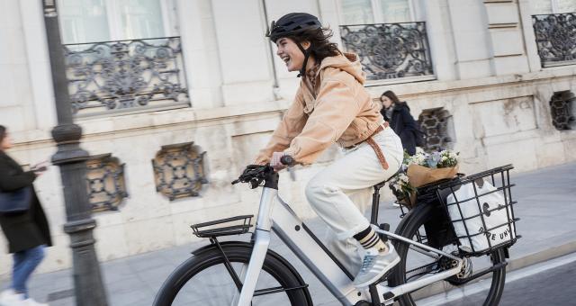A woman is riding a white e-bike in an urban setting. She is wearing white jeans and a peach sweater. She is laughing. The e-bike has a rear rack and panniers with flowers and shopping in