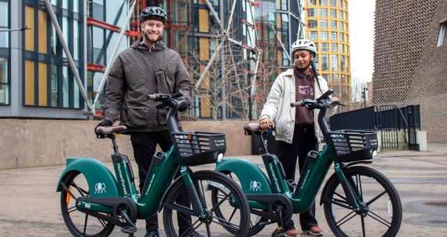 Two people are standing holding Human Forest e-bikes, green bikes with a black basket on the front and with the Human Forest logo. They are in an urban environment and wearing normal clothes and bike helmets.