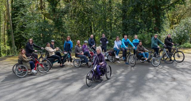 A group of adults in a park on a variety of non-standard cycles, tandems, e-assist cycles, handcycles and tricycles