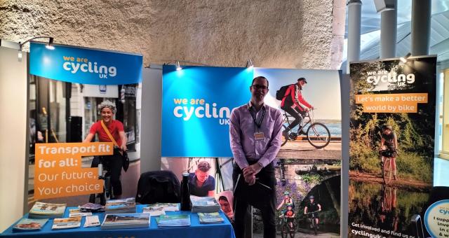 A man in shirt and trousers with a name tag round his neck and holding a clipboard. He is at an exhibition stand with Cycling UK branded posters and table