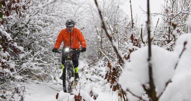 A man is cycling along a snow covered cycle track. Snow covered trees are on both sides of the track. The cyclist is wearing an orange coat and cycle helmet and riding a mountain bike