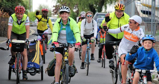 A mixed group of people is cycling along an urban road. There are different kinds of bikes and even a trailer with kids in. They are smiling and waving. Some are wearing hi-vis but most are in normal clothing.