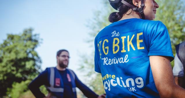 A woman wearing a blue T-shirt and cycle helmet is standing with her back to the camera. The T-shirt reads 'The Big Bike Revival; We are Cycling UK'