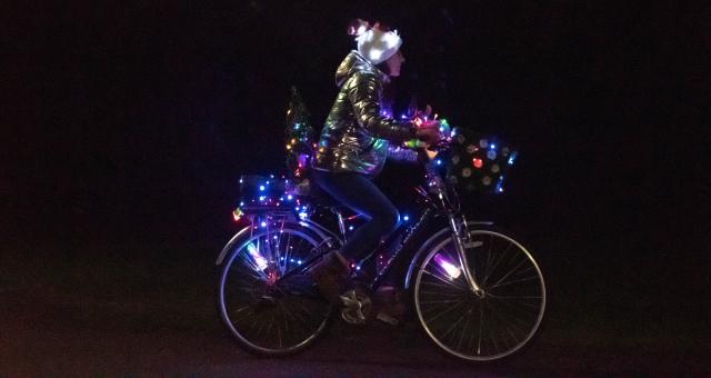 A female cyclist is riding a hybrid bike at night. It has a front basket and rear rack and there are loads of coloured lights all over it. She is wearing jeans, a gold jacket and a festive hat