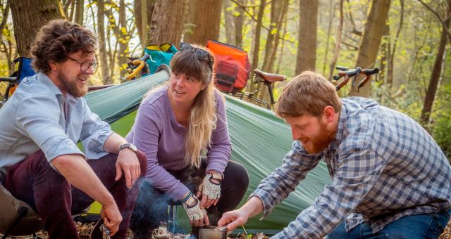 Three people are camping in the woods. They are making a hot drink. There's a tent in the background and you can see two packed gravel bikes leaning on trees