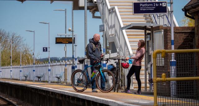 Two people are standing on a railway platform. They are both holding bikes.