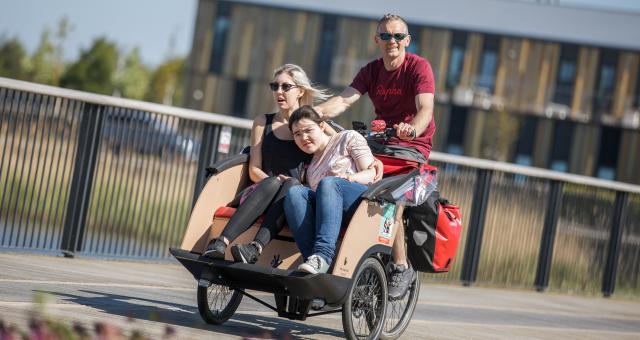 A man in sunglasses cycling an adaptive trike with seating in the front, carry two women as passengers