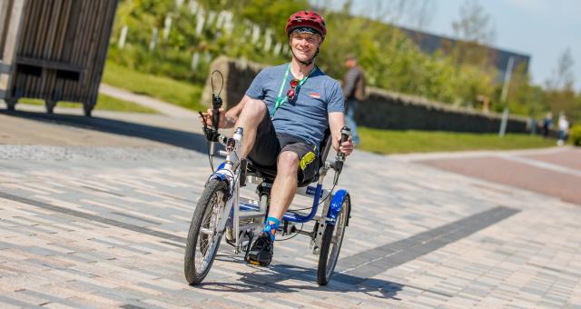 A man smiling at the camera as he cycles a recumbent trike out in the sun on a pedestrianised path