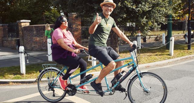 Two people are on a turquoise tandem cycling across a quiet junction with trees and a park in the background. The pilot is a man in shorts, T-shirt and sunhat. He's giving a thumbs up. The stoker is a woman in dark glasses. She's wearing leggings and a T-shirt.