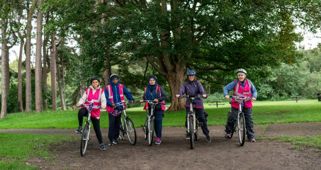 Five women are in a park standing with their bikes in front of a big tree. They are wearing a mix of cycling and normal clothing.