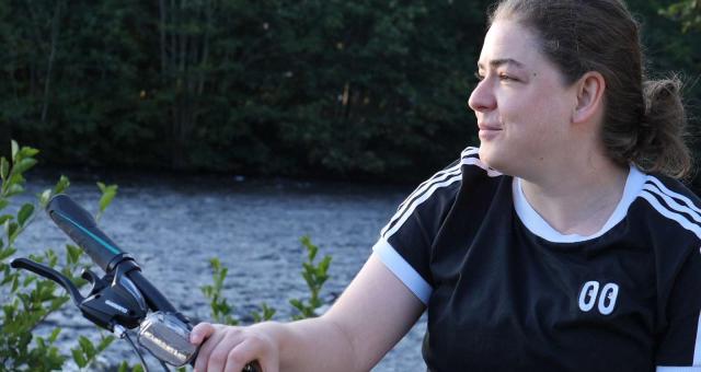 A woman in a navy blue t-shirt with white stripes sits on a bicycle by a lake while holding on to her handlebars