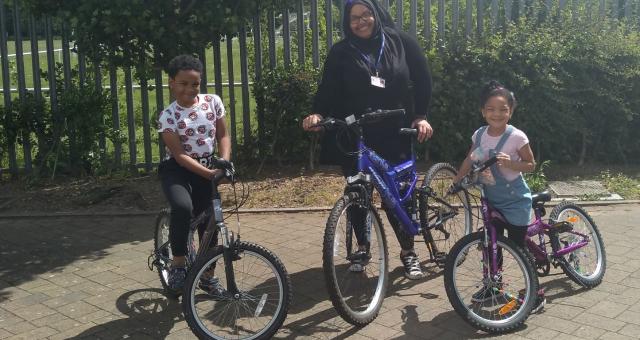 A woman and a boy and a girl pose with bicycles outside on a sunny day