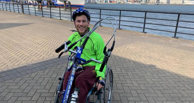 A man in a fluorescent green jacket sits in a handcycle at the waterside, behind him a ferry can be seen in the distance