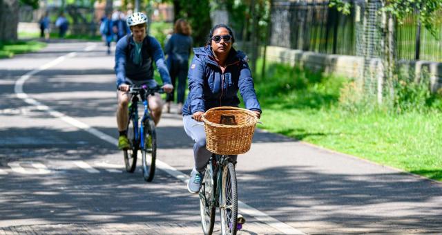 Two people are cycling on a dedicated cycle path alongside a pedestrian path. One is on a mountain bike, the other on a hybrid with a wicker basket on the front. They are both wearing normal clothes.