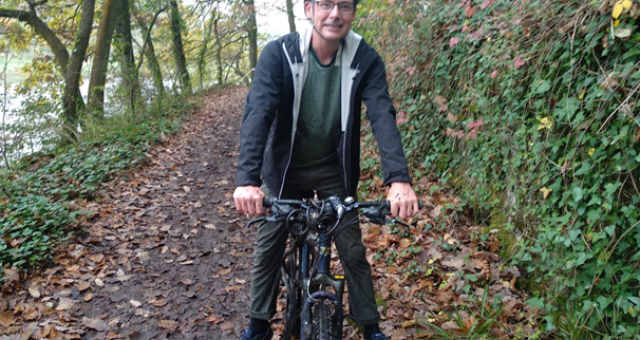 A middle-aged man wearing a green helmet sits astride a bicycle in a woodland setting