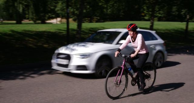A car accelerates past a female cyclist wearing a helmet