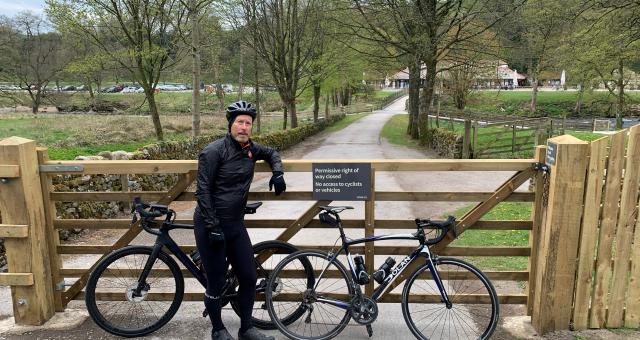 A cyclist dressed in black with two black bicycles stands in front of a barred wooden gate