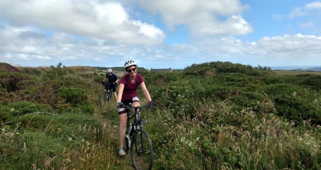 A young woman cycles towards us among a moorland vista followed by a young man
