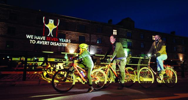 Two men and a young girl cycle on bikes in front of a lit up building