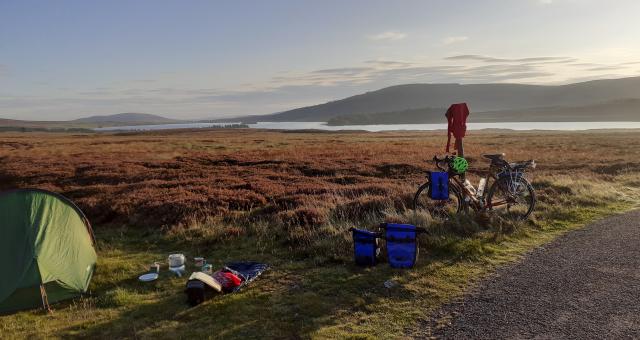 A bicycle is propped up against a signpost next to a pitched tent in front of a loch and mountains in the background