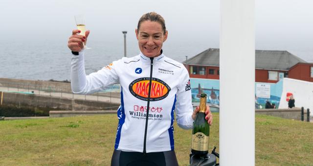 A woman holds up a glass of champagne as she stands underneath the John O' Groats sign in Scotland. She's wearing cycling jersey and bibs