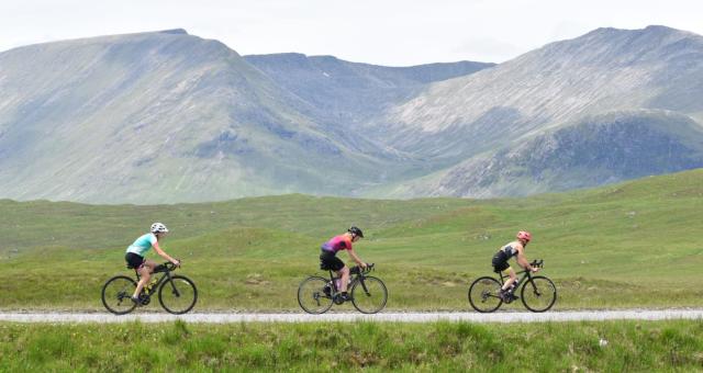 Three women road cycling in single file with a hazy mountain landscape in the background