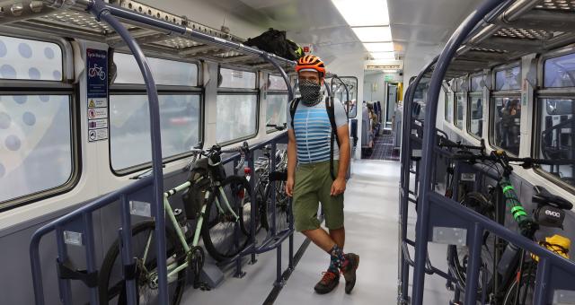 A man stands wearing a helmet. He‘s leaning against the pole in a train bicycle carriage in front of his gravel bike