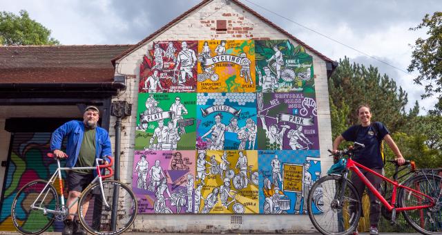 man and woman stand with bike each in front of colourful mural on gable end of old building in park