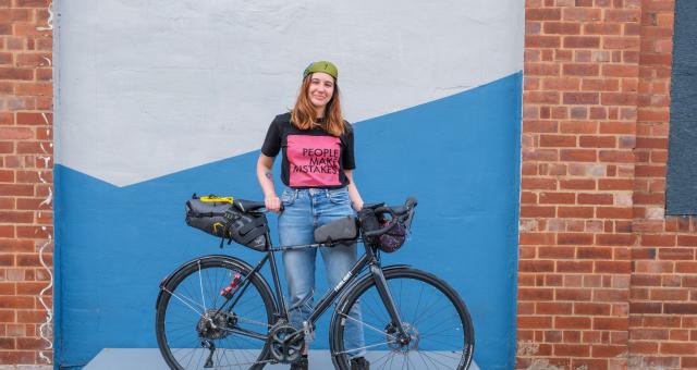 Adna stands smiling with her black bike. She is wearing a cap, black and pink slogan t-shirt and jeans.