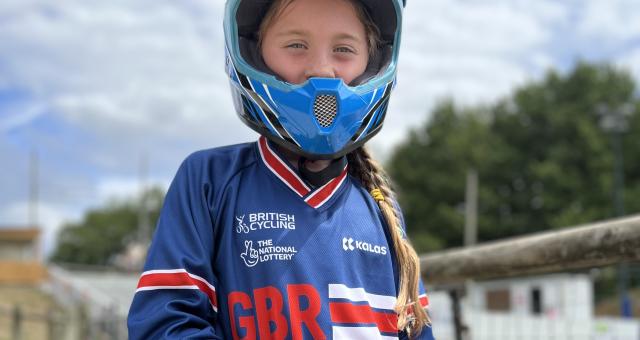Amelie stands with her BMX bike. She is wearing a BMX helmet, gloves, blue British Cycling jersey and green trousers.