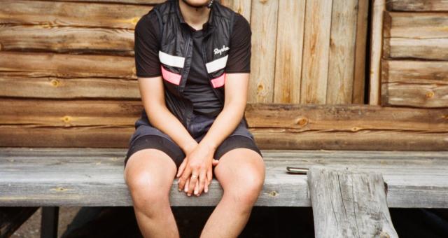Anna sits on a wooden veranda. She wears a helmet, black top, gilet, shorts and trainers.