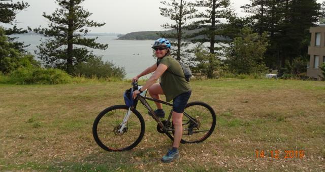Helen smiling on her bike. She wears a blue helmet, green top and dark shorts. There is a lake behind her.