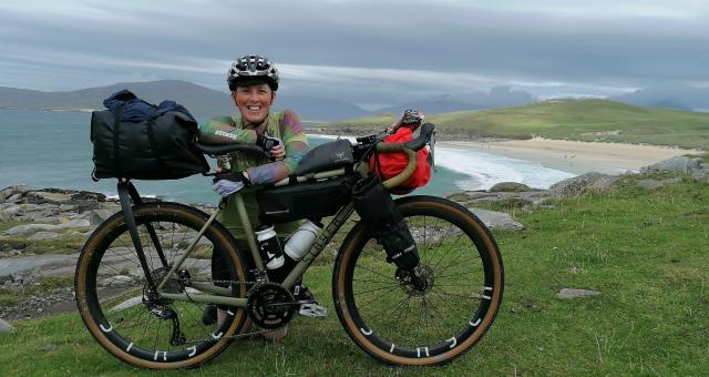 Iona kneels behind her green bike which is set up with bikepacking bags. She wears a helmet, green jersey, gloves and black shorts. A beach is in the background.