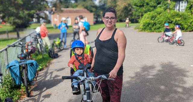 Kat stands to the right of her bike in a park. A small child is in a child’s seat at the back of the bike. Kat is wearing glasses, a black top and red patterned leggings.