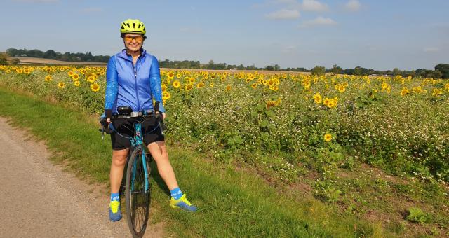 Marina stands next to a sunflower field with her blue bike. She wears sunglasses, a yellow helmet, blue jacket and black shorts.