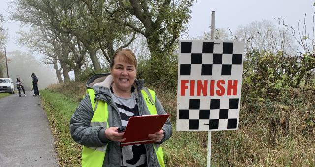 Sharon smiles to camera, holding a clipboard and stopwatch. She is standing at the side of the road at the finish line. She is wearing a grey winter coat and hi-vis vest.