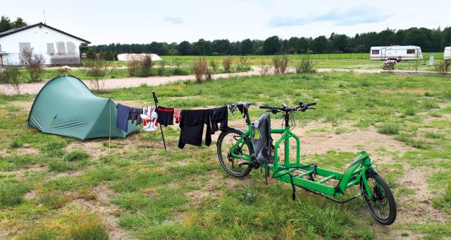 A tent sits on a grassy campsite, with a washing line of cycling clothes hung between it and a bright green cargo bike