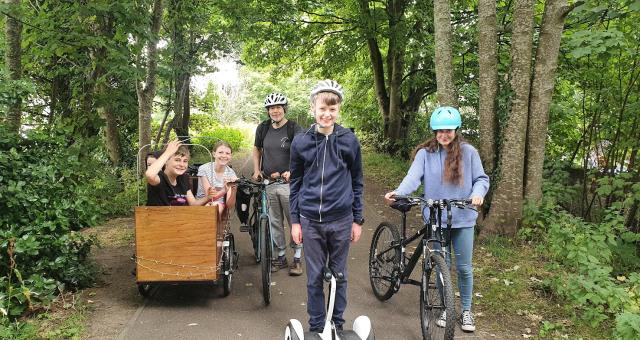 Three teenage children sit in a cargo bike next to a man standing with his bicycle. A young boy stands in the foreground with a segway and a young lady stands to his left holding a mountain bike.