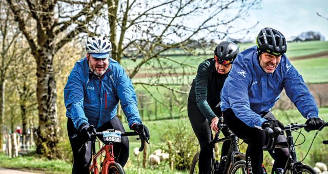 Three men cycle along a road with green fields in the background.