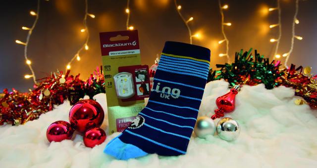 A pair of blue stripy socks display the Cycling UK logo. They lay next to a front bicycle light and are surrounded by christmas baubles and fairy lights