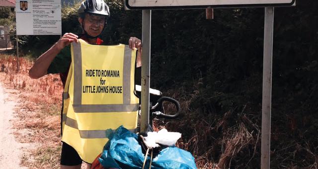 A man stands in front of a road sign that says 'Cisnadioara Michelsberg'. He is holding up a high-vis tabard that bears the words 'Ride to Romania for Little John's House'. He is wearing a helmet and his bicycle leans next to the road sign.
