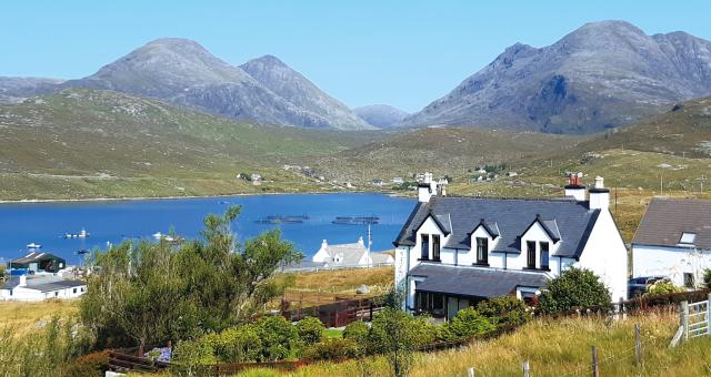 A white cottage sits in a green field in front of a lake and mountains