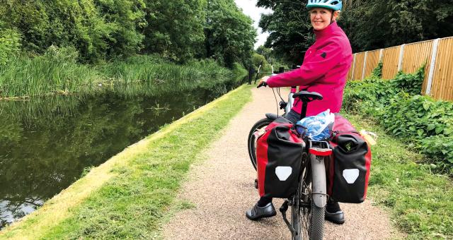 A woman sat on a bicycle laden with full rear panniers turns to look at the camera. She has paused on a canal towpath
