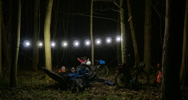 Three men sit in a hammock in some woods at night lit up by a string of fairy lights