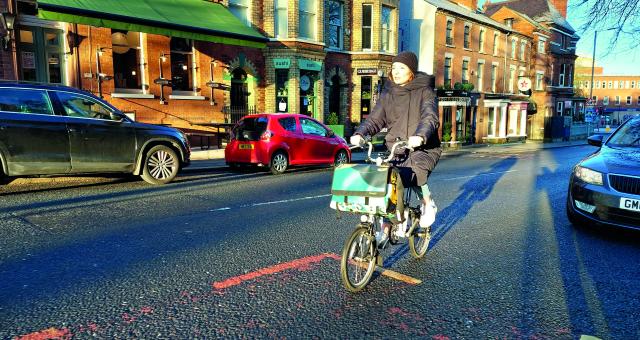 A woman cycles along a main road lined with shops and parked cars. She is wearing casual winter clothes